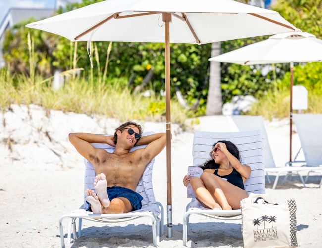 A couple relaxes on beach chairs under a white umbrella, with a beach bag nearby and vegetation in the background, ending the sentence.