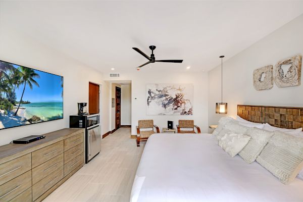 The image shows a modern, well-decorated hotel room with a large bed, flat-screen TV, ceiling fan, mini-fridge, and stylish artwork on the walls.