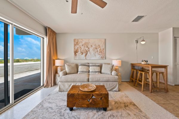 A cozy living room with a large window, a beige sofa, a wooden coffee table, a painting, lamps, and a small bar area with stools, all well-lit.