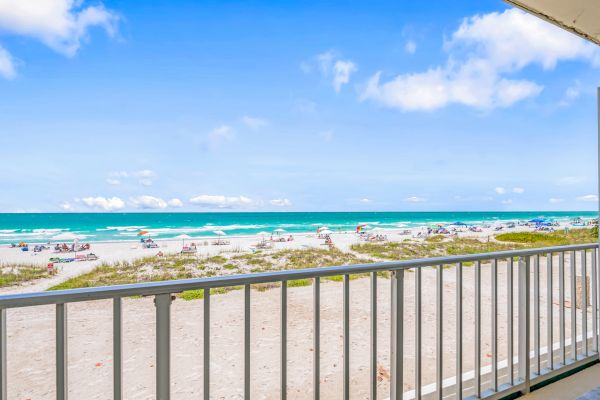 A balcony view of a sandy beach with people, umbrellas, and the ocean under a bright blue sky with some clouds, ending the sentence.