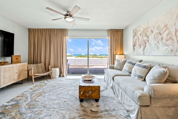 A modern living room with a large sofa, patterned rug, wooden furniture, and sliding doors opening to a beach view with blue skies.