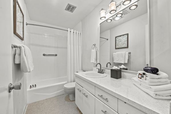 This image shows a clean, white bathroom with a vanity, mirror, bathtub, shower curtain, towels, and a well-lit environment.