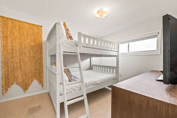 The image shows a small bedroom with a white bunk bed, a dresser with a TV, a window, and a wall hanging made of small yellowish items.