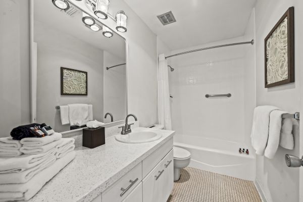 A modern bathroom with a large mirror, sink, toilet, and shower-tub combo. Towels, a wall-mounted artwork, and vanity lights are visible.