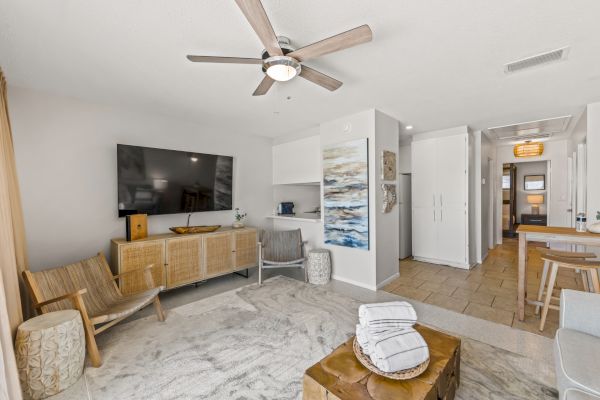 A modern living room with a ceiling fan, wall-mounted TV, wooden furniture, and coastal decor. The image includes a hallway and a small dining area.