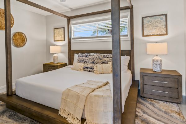 A cozy bedroom with a wooden four-poster bed, two bedside tables with lamps, and decorative wall art. The room has a serene, neutral palette.