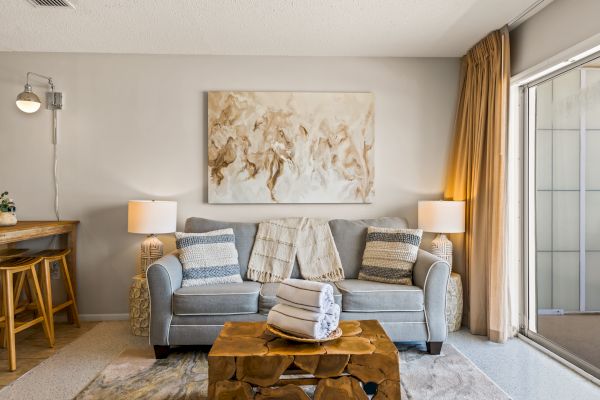 A cozy living room with a gray sofa, abstract art, two lamps, a unique wooden coffee table, rolled towels, and a small bar area to the left, ending the sentence.