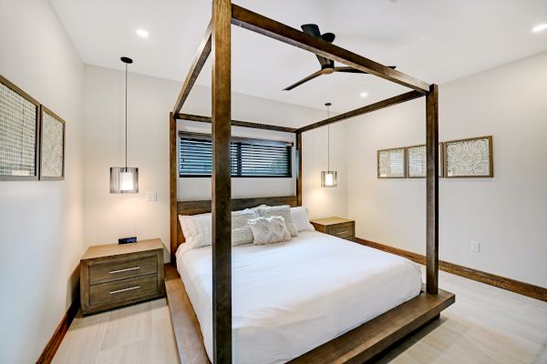 A modern bedroom with a wooden canopy bed, white bedding, and nightstands. It features hanging lights, framed art, and a ceiling fan on the light wood floor.