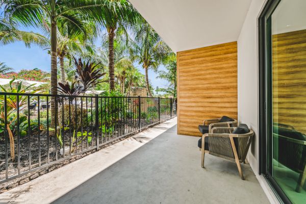 A sunny outdoor patio with two chairs, a railing, tropical plants, and a sliding glass door reflecting the greenery.