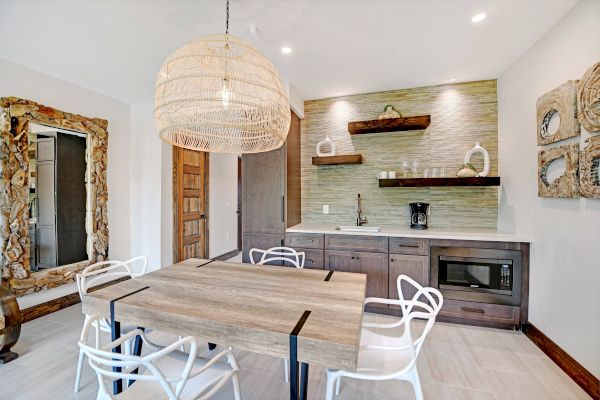 A modern dining room with a wooden table, white chairs, a large woven pendant light, wall art, and a kitchenette with shelves and a microwave.