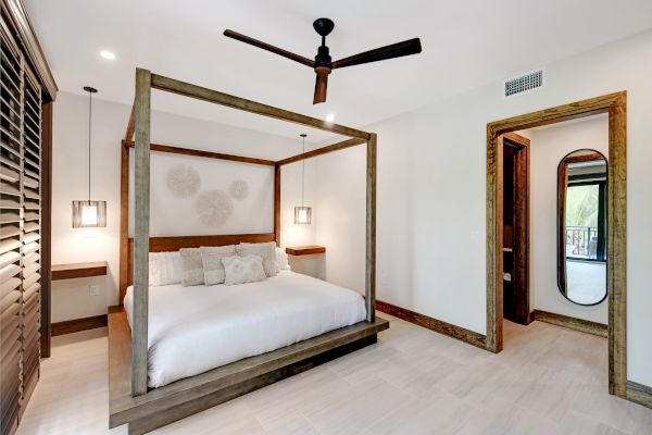 A minimalist bedroom with a canopy bed, modern ceiling fan, wall art, bedside tables with lamps, and a doorway leading to another room.