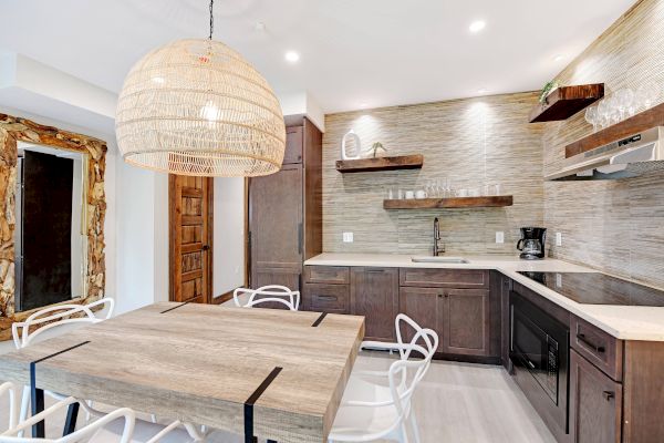 Modern kitchen with wooden cabinets, large pendant light, dining table, white chairs, shelves, and built-in appliances, featuring a rustic door.