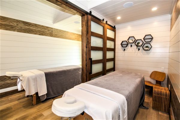 A modern, serene massage room with a massage table, wooden decor, shelves with small plants, and soft lighting in a cozy environment.