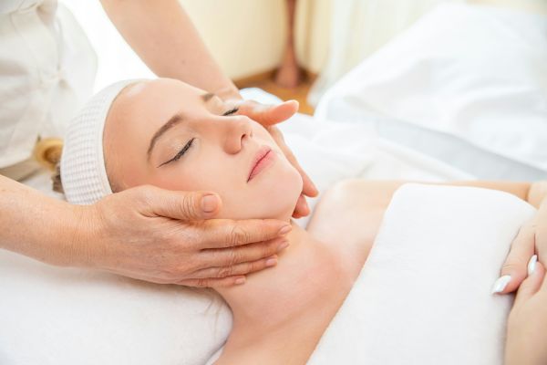 A woman is lying down with her eyes closed, receiving a facial massage from a person’s hands. She wears a headband and is covered with a white towel.