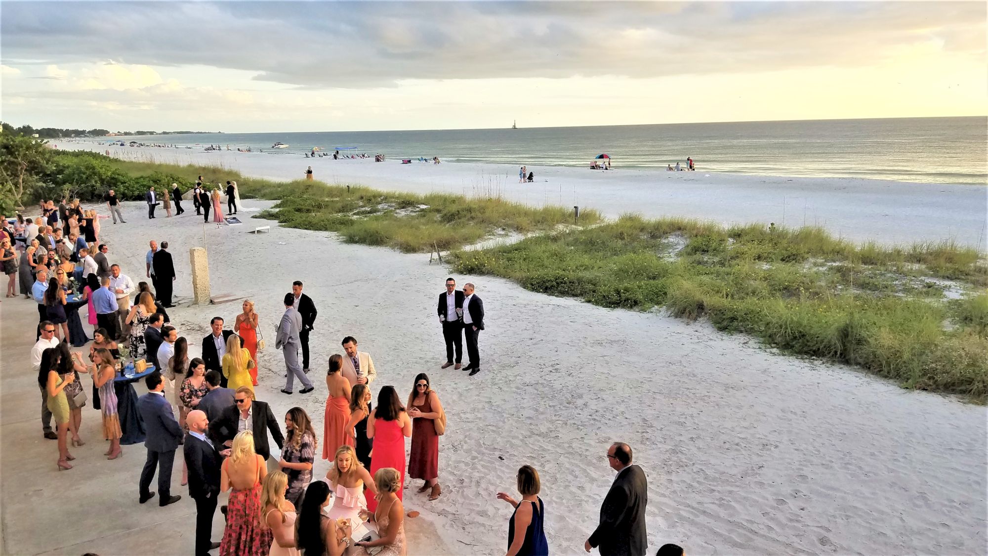 A gathering of people dressed formally on a sandy beach near the ocean. Some are standing in groups and others are walking or sitting.