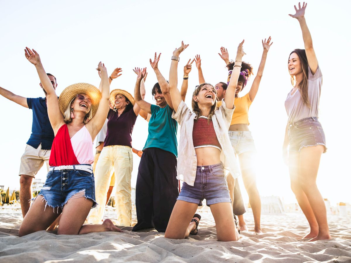 A group of people are enjoying themselves on a sandy beach, raising their hands in the air, smiling, and wearing casual summer clothes.