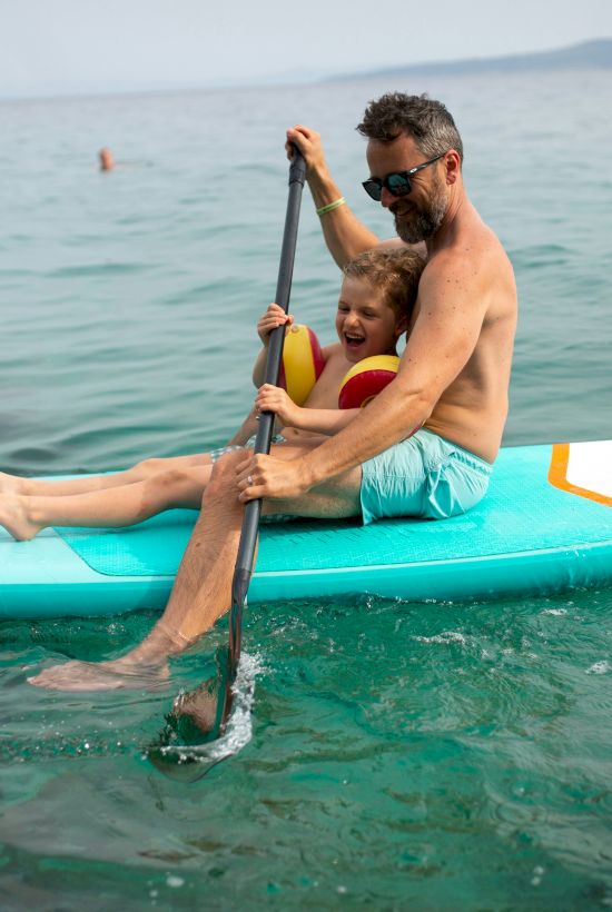 A man and a child are paddling together on a paddleboard in calm, clear waters, with distant land visible in the background.