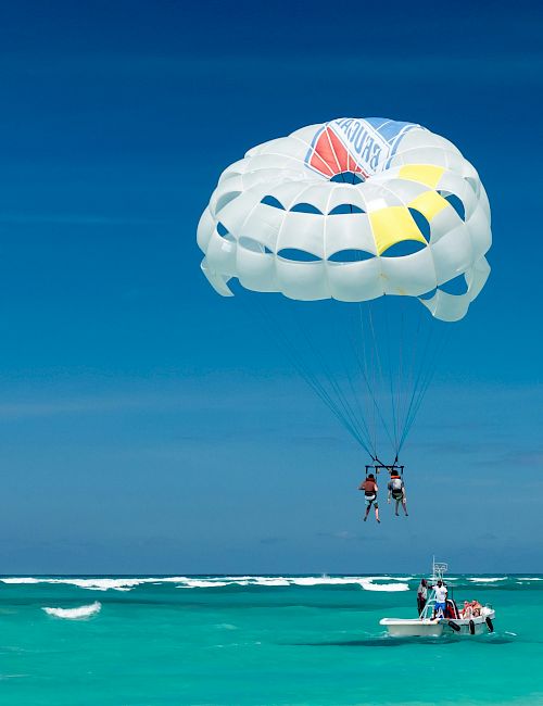 Two people are parasailing over the turquoise ocean, attached to a parachute and pulled by a boat on a clear, sunny day.