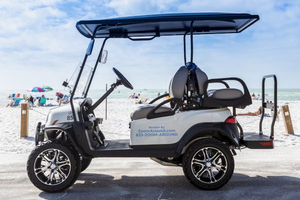 The image shows a golf cart parked on a paved area near a beach. Several people and umbrellas are visible on the sandy beach in the background.