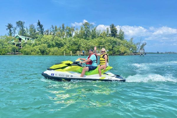 Two people riding a jet ski on a sunny day, with a lush island and trees in the background across a body of water.