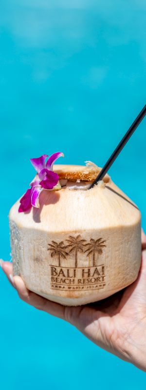 A hand holds a coconut drink adorned with a flower and straw against a blue water background. The coconut is labeled 