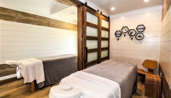 The image shows a serene massage room with a table, cozy linens, wooden decor, soft lighting, and wall shelves holding small plants, creating a relaxing vibe.