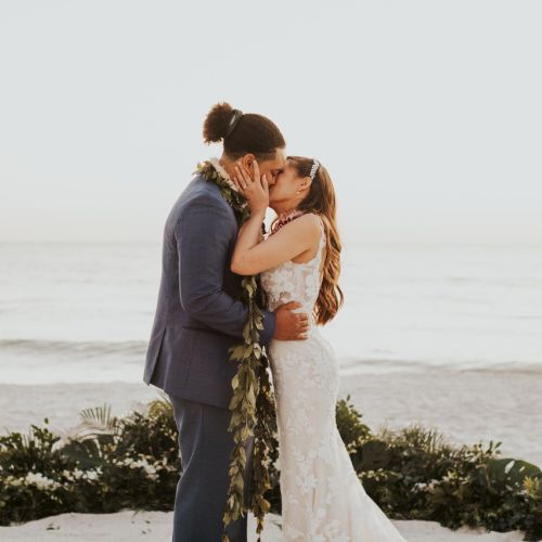 A couple shares a kiss in a beachside wedding ceremony, surrounded by greenery and ocean waves in the background.