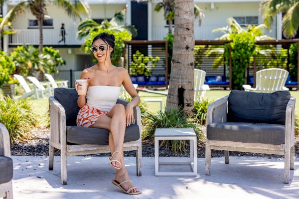 A woman sits outdoors on a patio chair, wearing sunglasses and holding a drink, with greenery and more seating visible in the background.